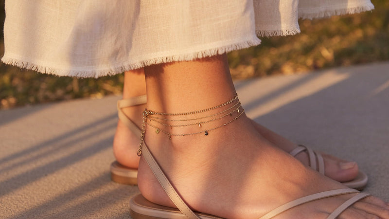Layered gold anklet on ankle with open sandals, warm sunlight editorial product photo